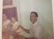 Two small children sit on a window sill with their smiling father next to them.