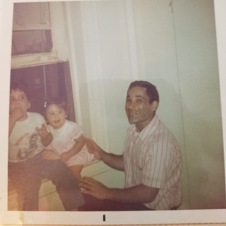 Two small children sit on a window sill with their smiling father next to them.