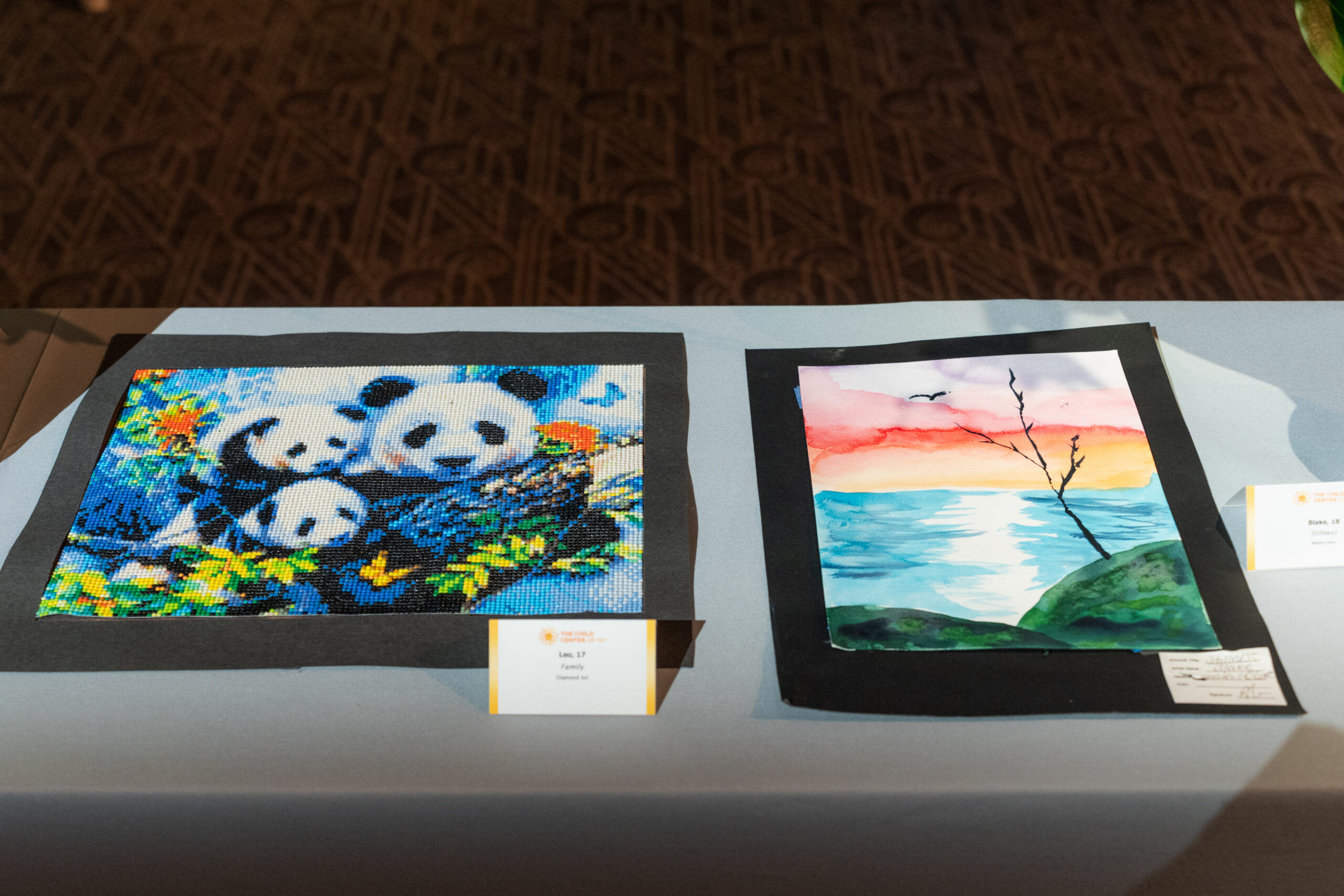 Needlework featuring pandas and a landscape paining are displayed on a table.