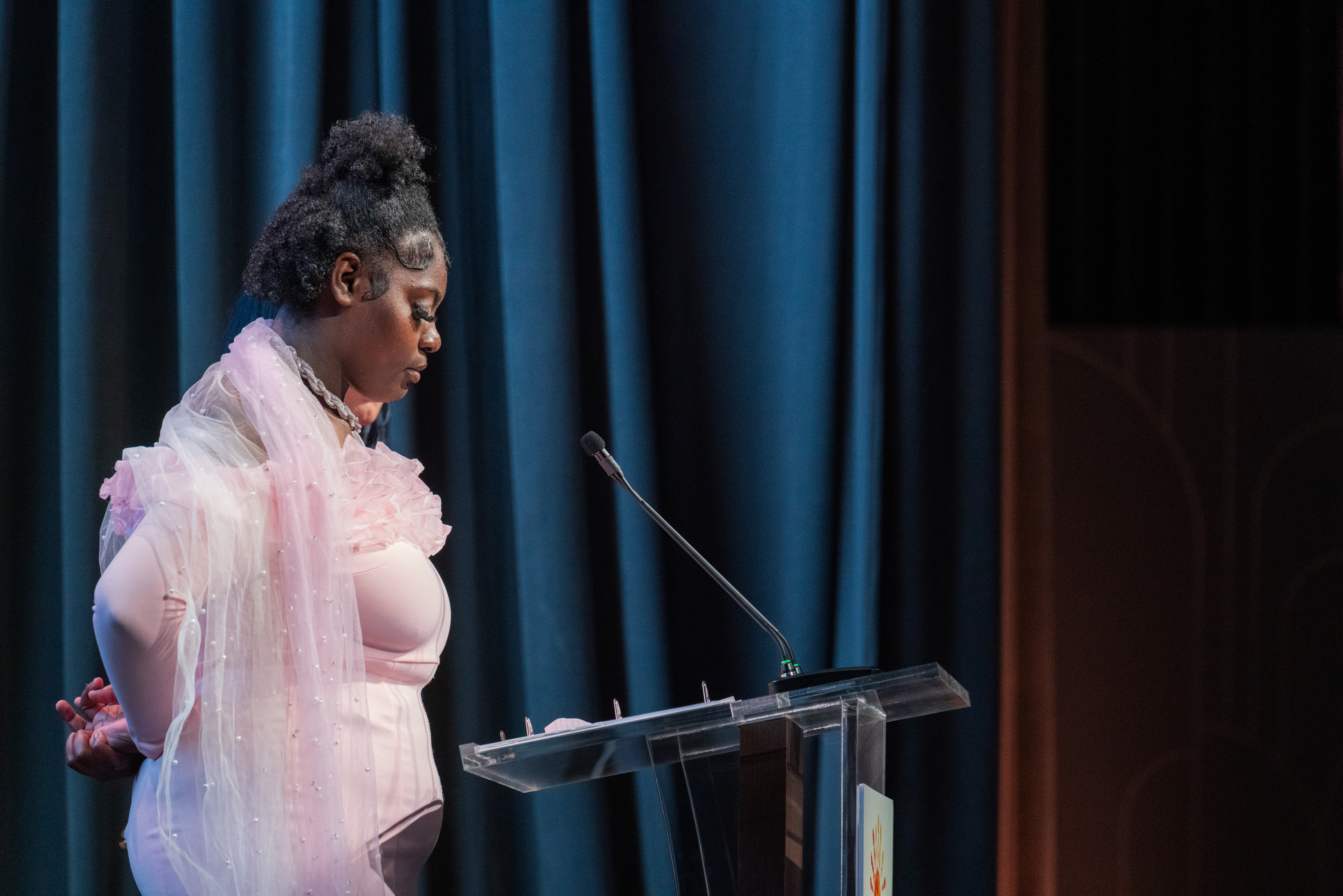 A young woman in a light pink dress stands at a podium and microphone at Evening Celebrating Resilience, the child center gala.