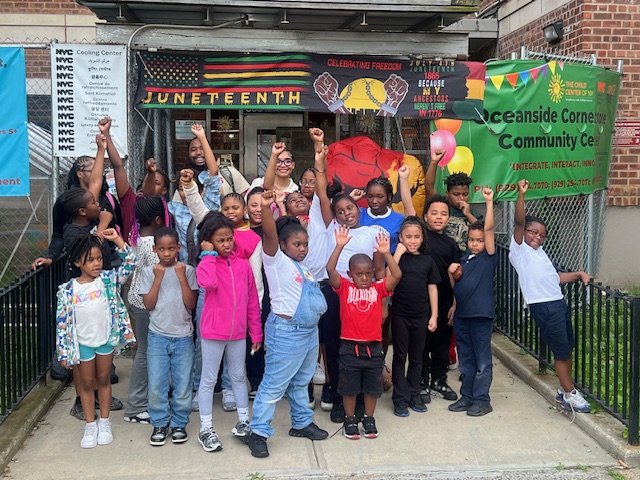 Youth and staff gather outside Oceanside Cornerstone Community Center during their Juneteenth celebration. Some are raising their fists. Banners are behind them.