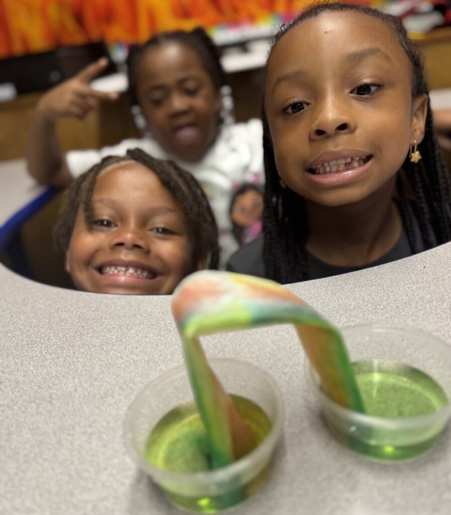 Three early elementary school age children watch in wonder as a folded napkin with its ends in cups of colored water turn into a "walking rainbow" 