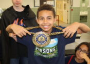 A smiling eighth grade boy wears a medal on a gold chain; the medal says, "Parsons Summer Rising Winner."